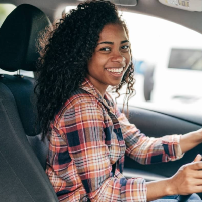 Smiling young woman with curly hair wearing a plaid shirt, sitting in the driver's seat of a car