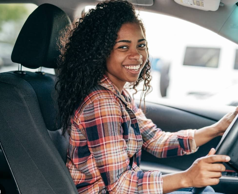 Smiling young woman with curly hair wearing a plaid shirt, sitting in the driver's seat of a car